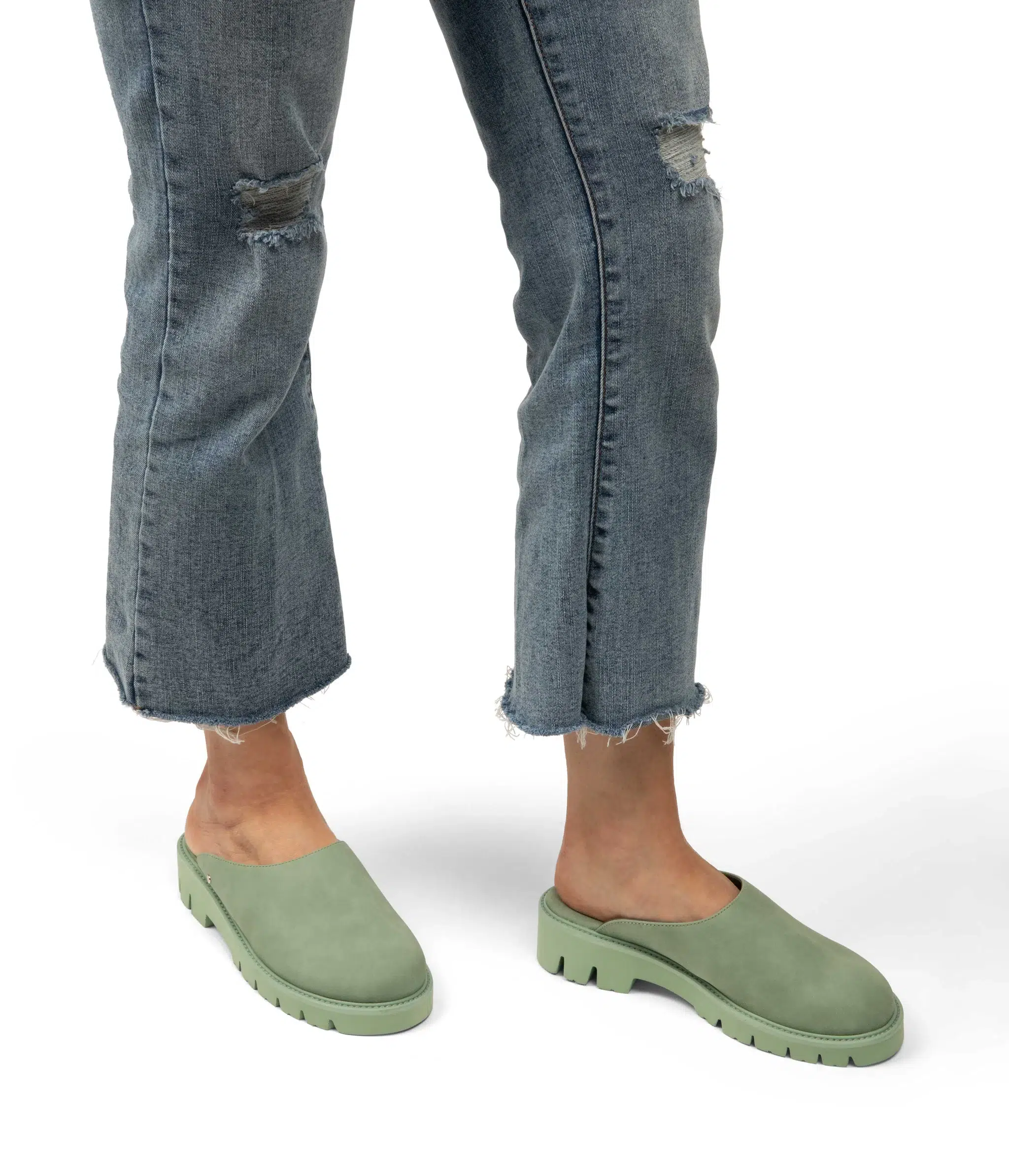 Person wearing frayed jeans and green slip-on clogs stands on a white background.