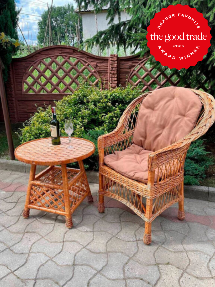 Wicker chair with a cushion and a round wicker table on a patio. A bottle of wine and two glasses are on the table. Wooden trellis fence and greenery in the background. "Reader Favorite" badge on top right.