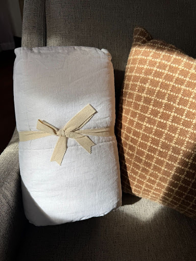 A rolled white comforter tied with a beige ribbon rests on a gray chair next to a brown patterned pillow.