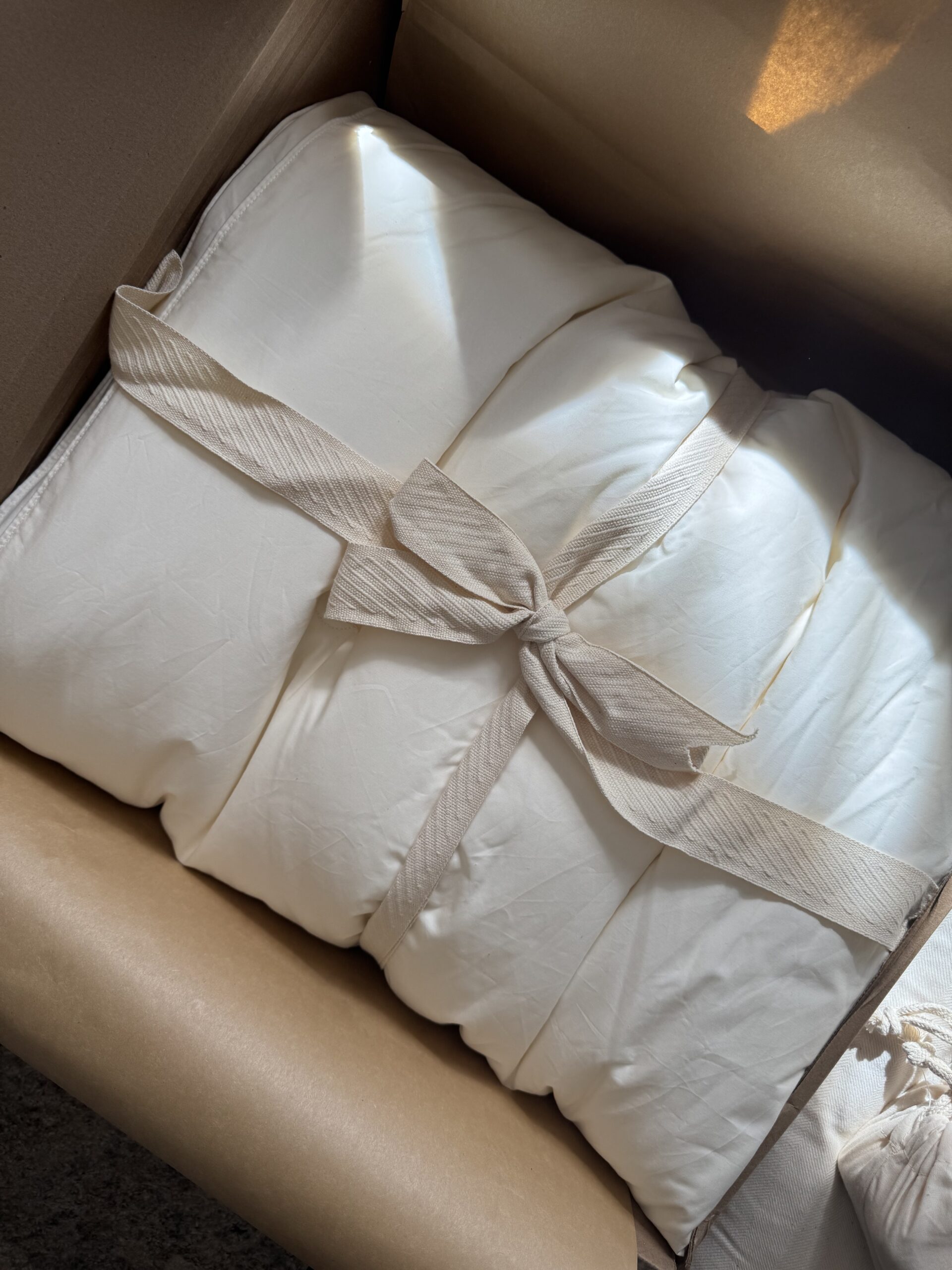 A neatly folded white comforter tied with a beige ribbon, placed inside a cardboard box.