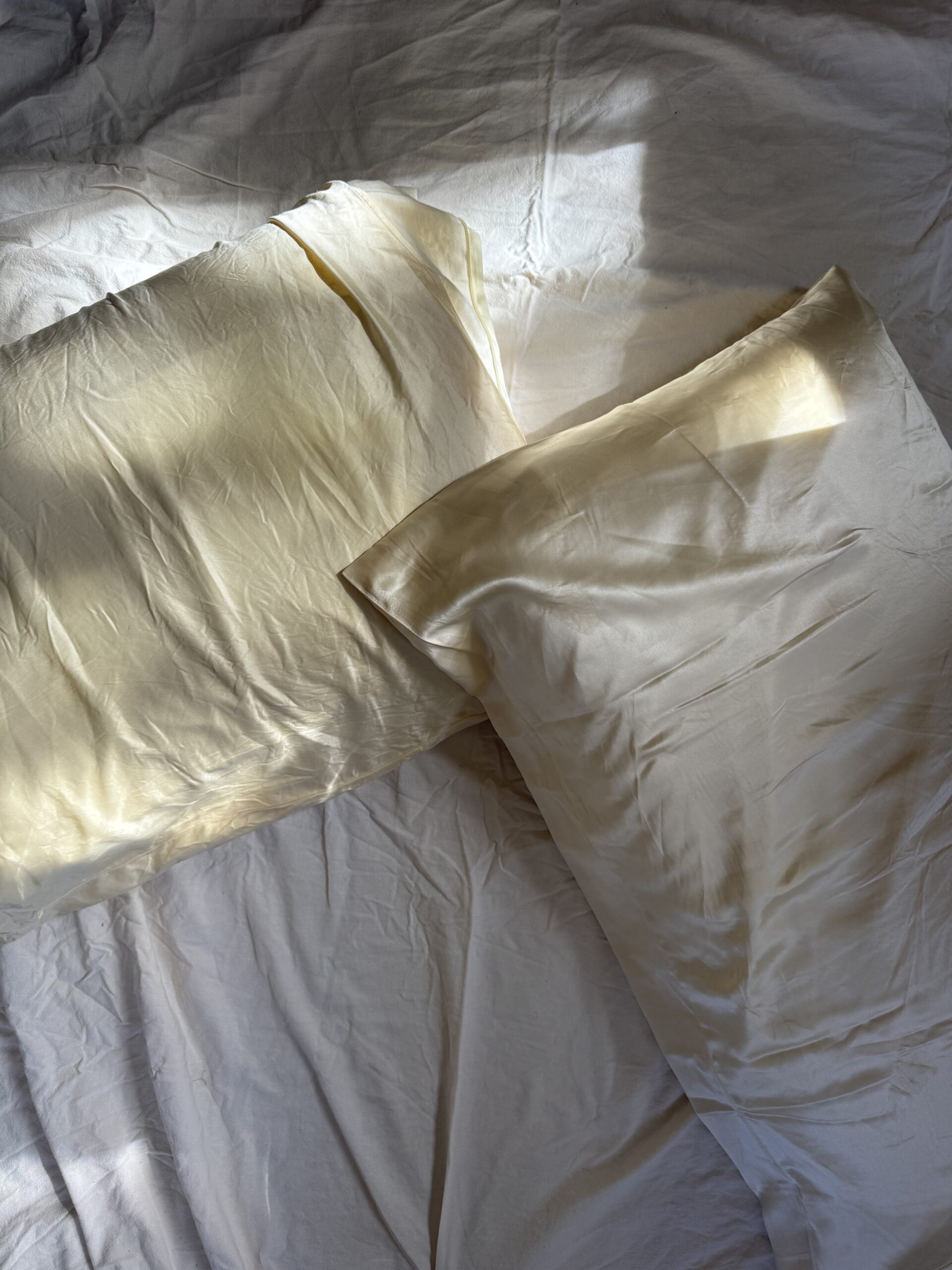 Two cream-colored satin pillows on a slightly wrinkled white bedsheet, with sunlight casting gentle shadows across the fabric.