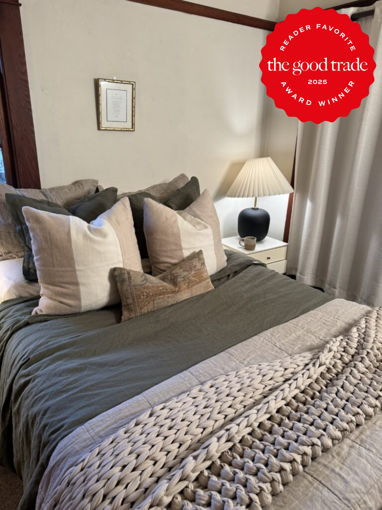 Cozy bedroom with a neatly made bed, large pillows, textured throw, and a bedside table with a lamp. A framed certificate is on the wall. "The Good Trade 2025 Reader Favorite" badge is visible.
