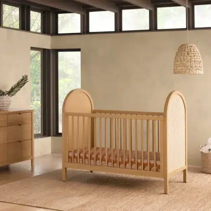 A wooden crib with a striped mattress is placed in a modern room with large windows, a dresser, and a woven light fixture.