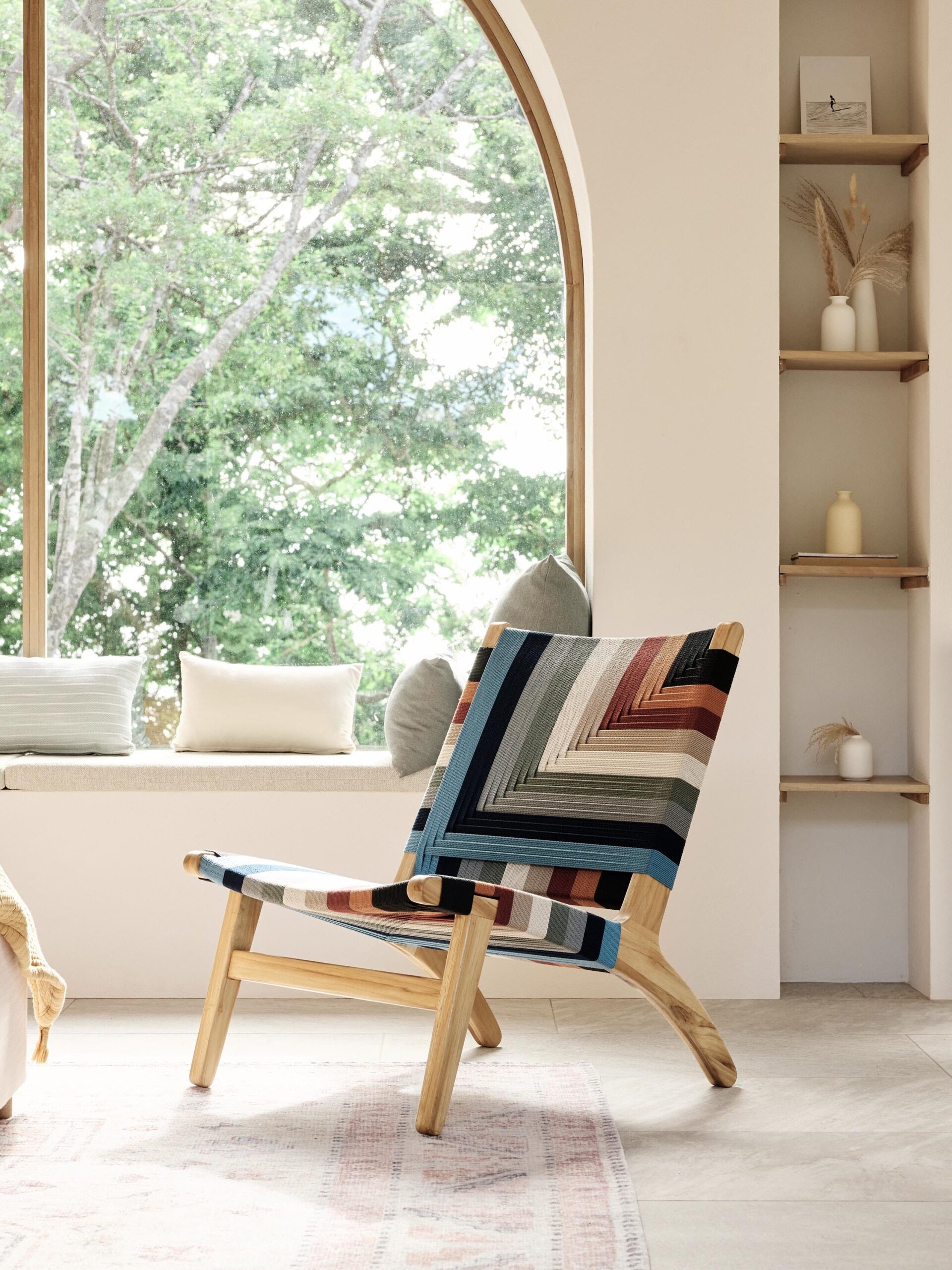 A wooden chair with a colorful geometric fabric sits in a softly lit room next to a large arched window. The window overlooks trees, and a bed with cushions is nearby.