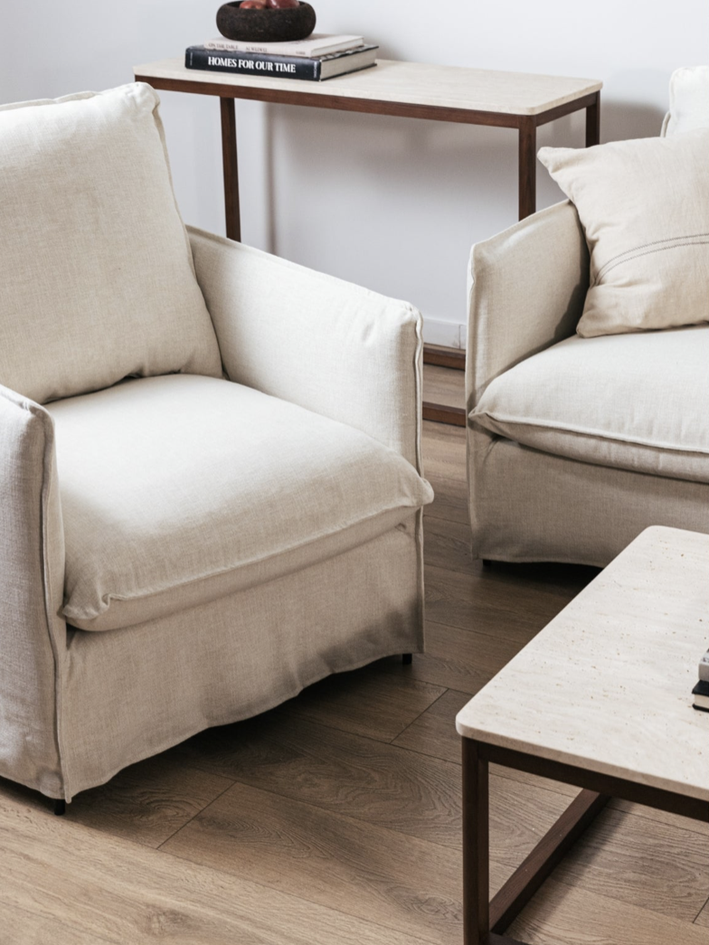 Two beige armchairs with plush cushions face a wooden coffee table. A console table with books and decor is in the background, set on wooden flooring.