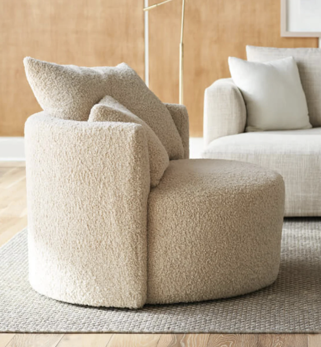 A round, beige boucle swivel chair on a gray rug, with a beige sofa and a floor lamp in the background.