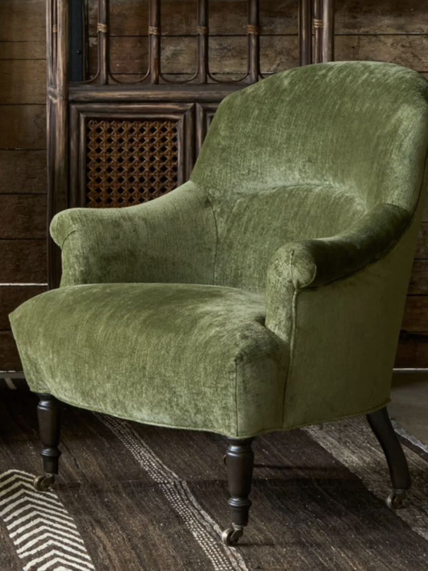 A green upholstered armchair with curved arms and black wooden legs on a dark patterned rug, set against a rustic wooden wall.