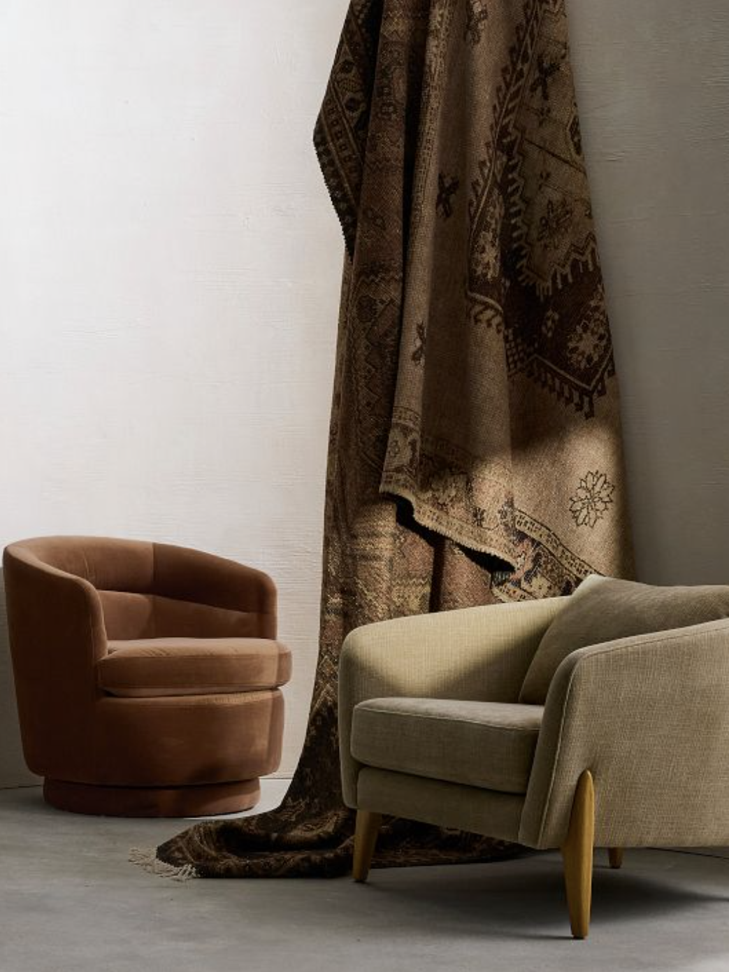A brown upholstered chair and a tan low-profile chair with wooden legs sit near a patterned rug draped over a wall.