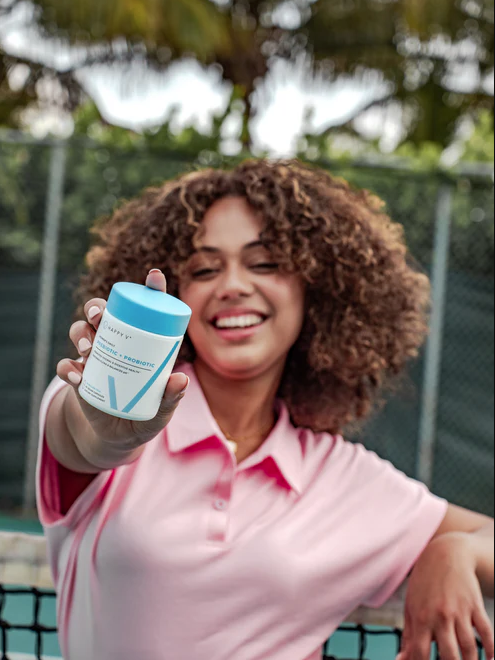 Person in a pink shirt holding a fitness supplement bottle outdoors, with trees and a fence in the background.