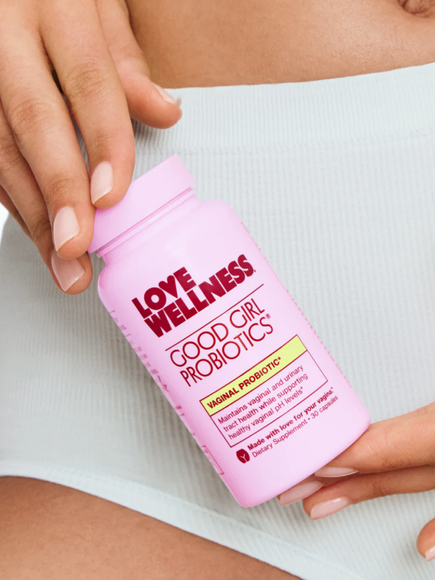 A person holding a pink bottle of "Love Wellness Good Girl Probiotics" against their waist, wearing light-colored shorts.