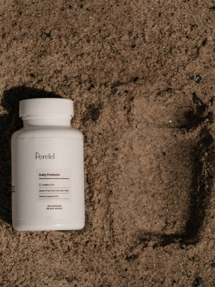 A bottle of Perelel Daily Probiotic is placed on sand next to its imprint.