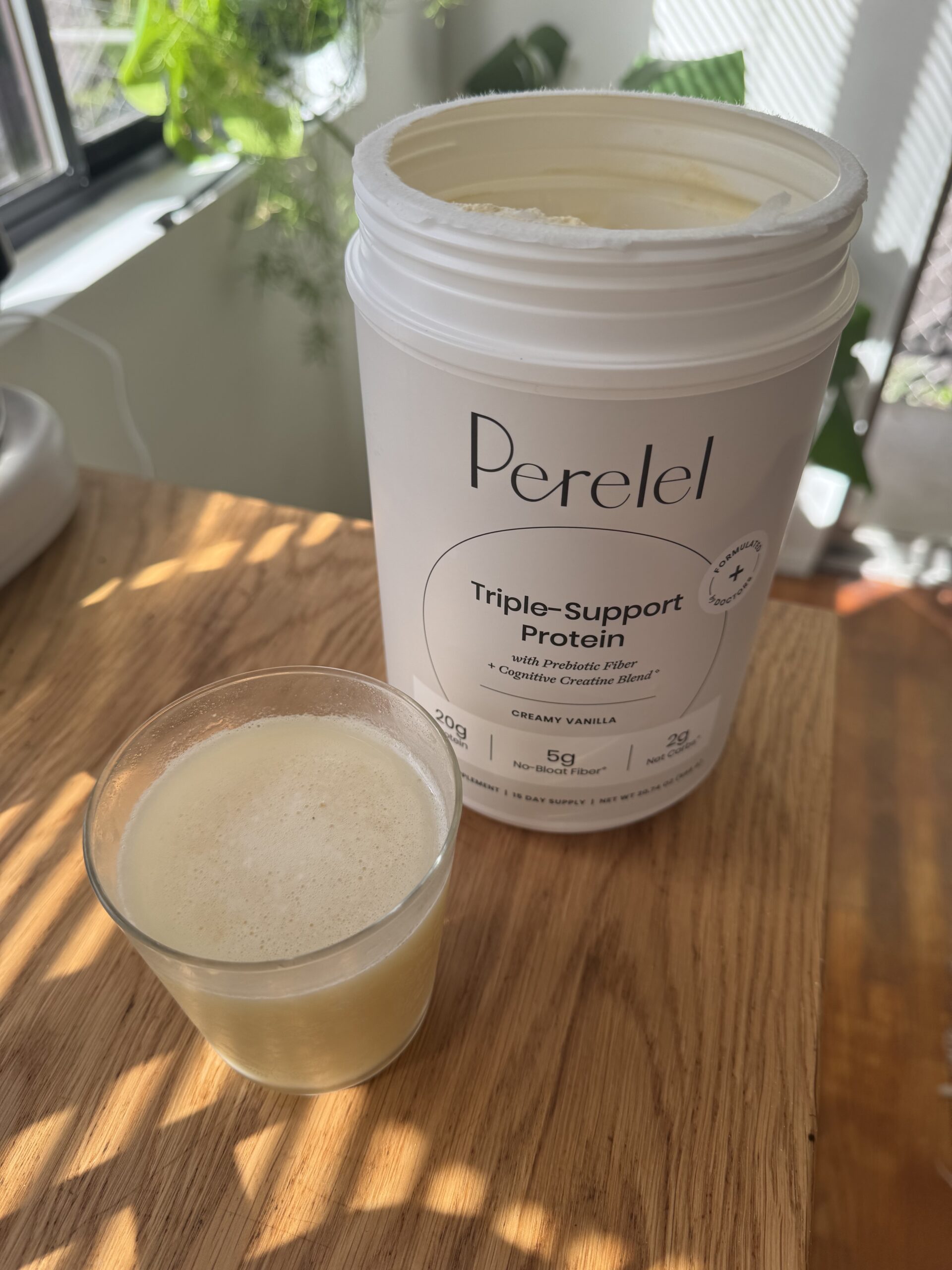 A jar of Perelel Triple-Support Protein in creamy vanilla flavor on a wooden table, next to a glass containing a protein shake. Plants and sunlight are visible in the background.
