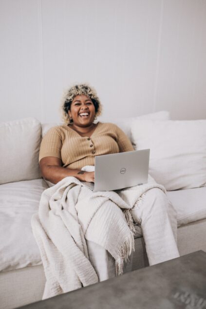 A person sits on a white couch with a beige knit blanket, using a silver Dell laptop to browse sustainable brands, and smiling.