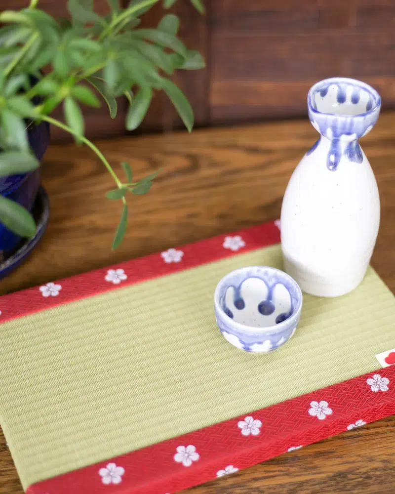 A white and blue ceramic sake bottle and cup sit on a green and red floral mat beside a potted plant on a wooden surface.