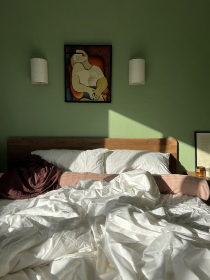 Unmade bed with white sheets, a brown blanket, and pillows. Two wall-mounted lamps and a framed abstract art piece hang on a green wall above the headboard. Sunlight casts shadows across the bed.