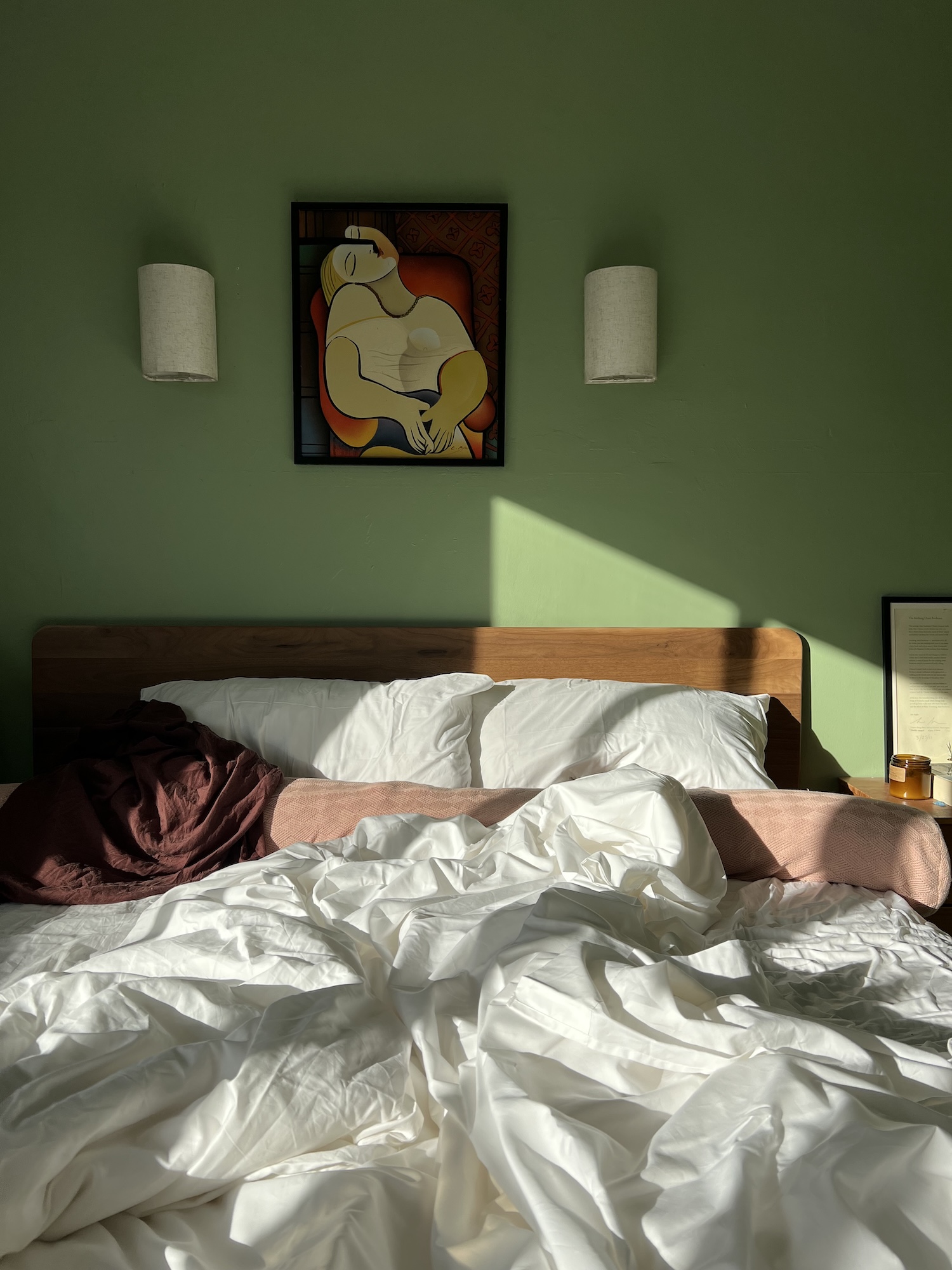 Unmade bed with white sheets, a brown blanket, and pillows. Two wall-mounted lamps and a framed abstract art piece hang on a green wall above the headboard. Sunlight casts shadows across the bed.