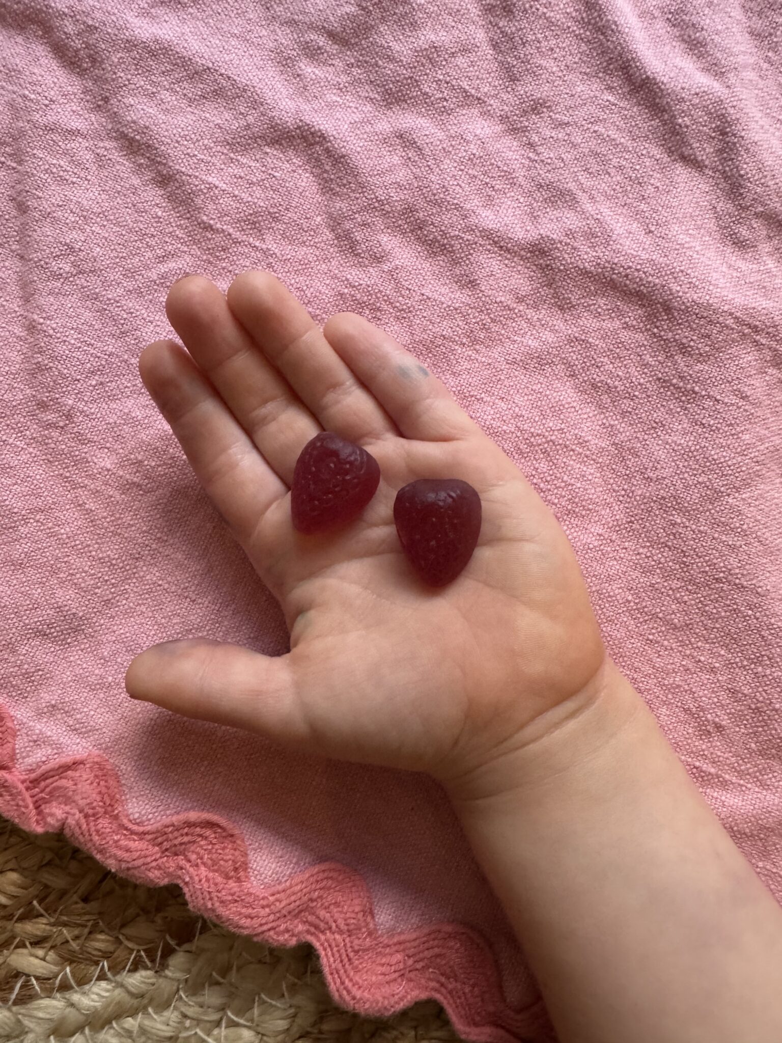 A child’s hand holding two heart-shaped red gummies, with a pink textured blanket in the background.