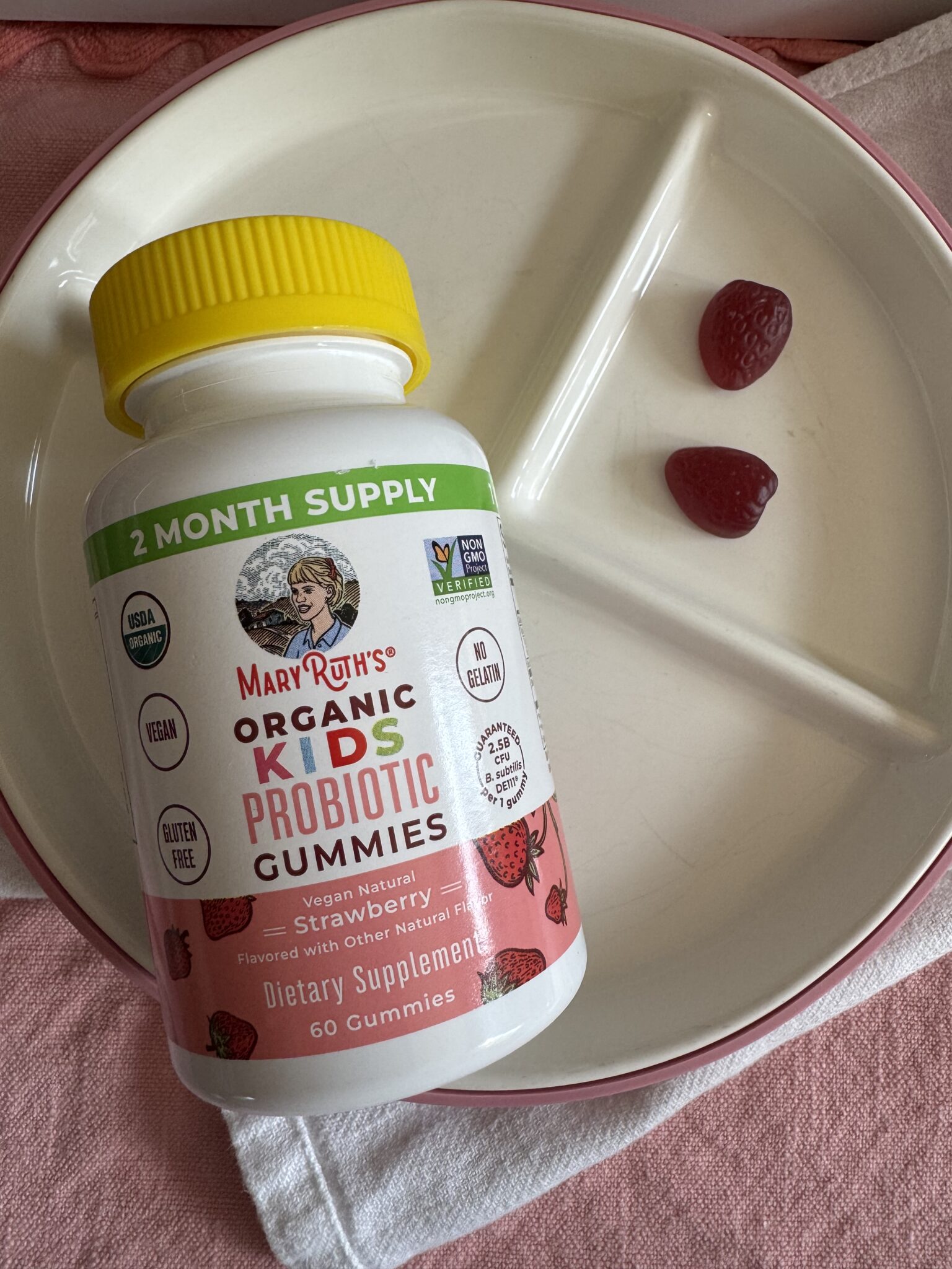 A bottle of MaryRuth's Organic Kids Probiotic Gummies lies on a plate next to two red gummy supplements shaped like strawberries.