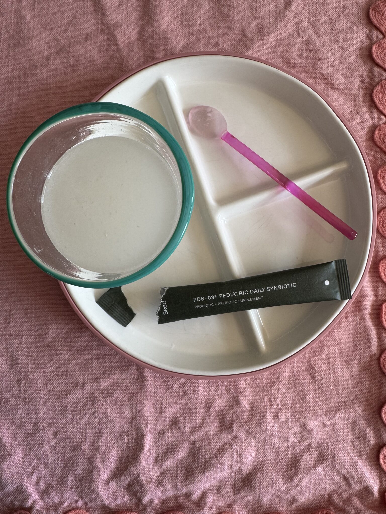 A divided plate holds a glass bowl with white liquid, a pink spoon, and an opened packet labeled "PDS-08 Pediatric Daily Synbiotic" on a pink cloth background.