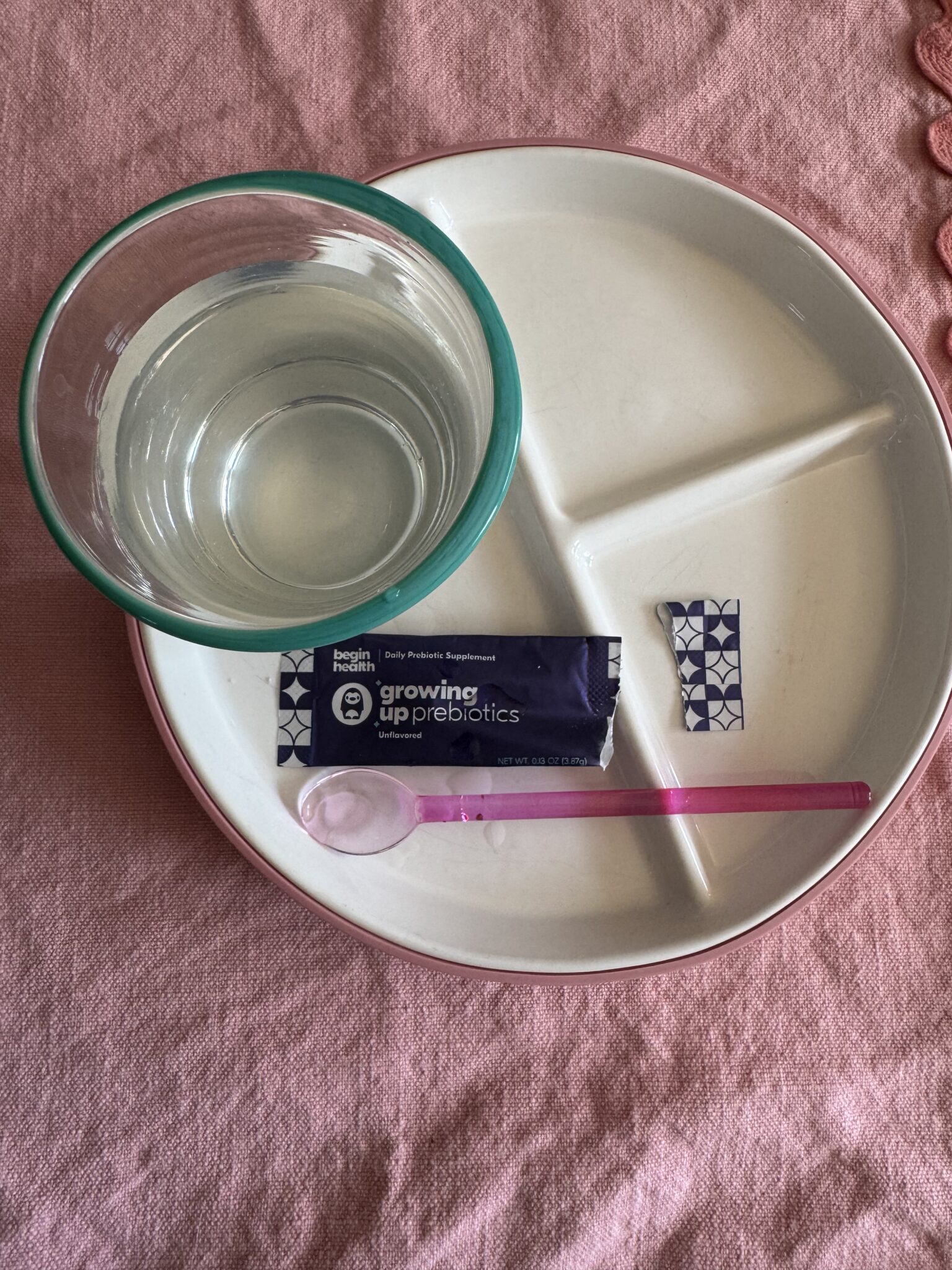 A divided plate holds a glass of water, a packet labeled "growing up prebiotics," a small square packet, and a pink plastic spoon on a pink surface.
