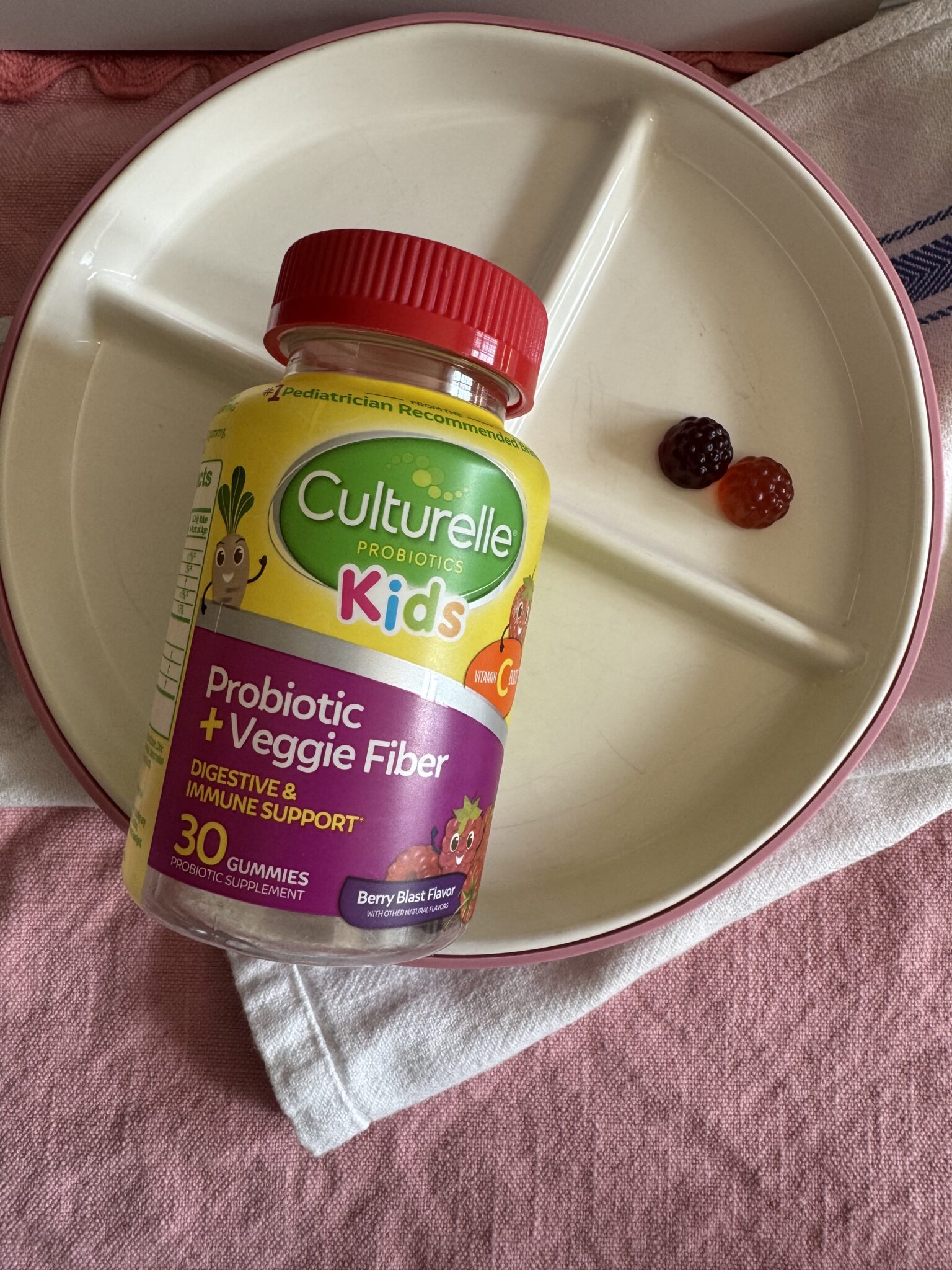 A bottle of Culturelle Kids Probiotic + Veggie Fiber gummies on a divided plate with two berry-shaped gummies, on a pink and white cloth.