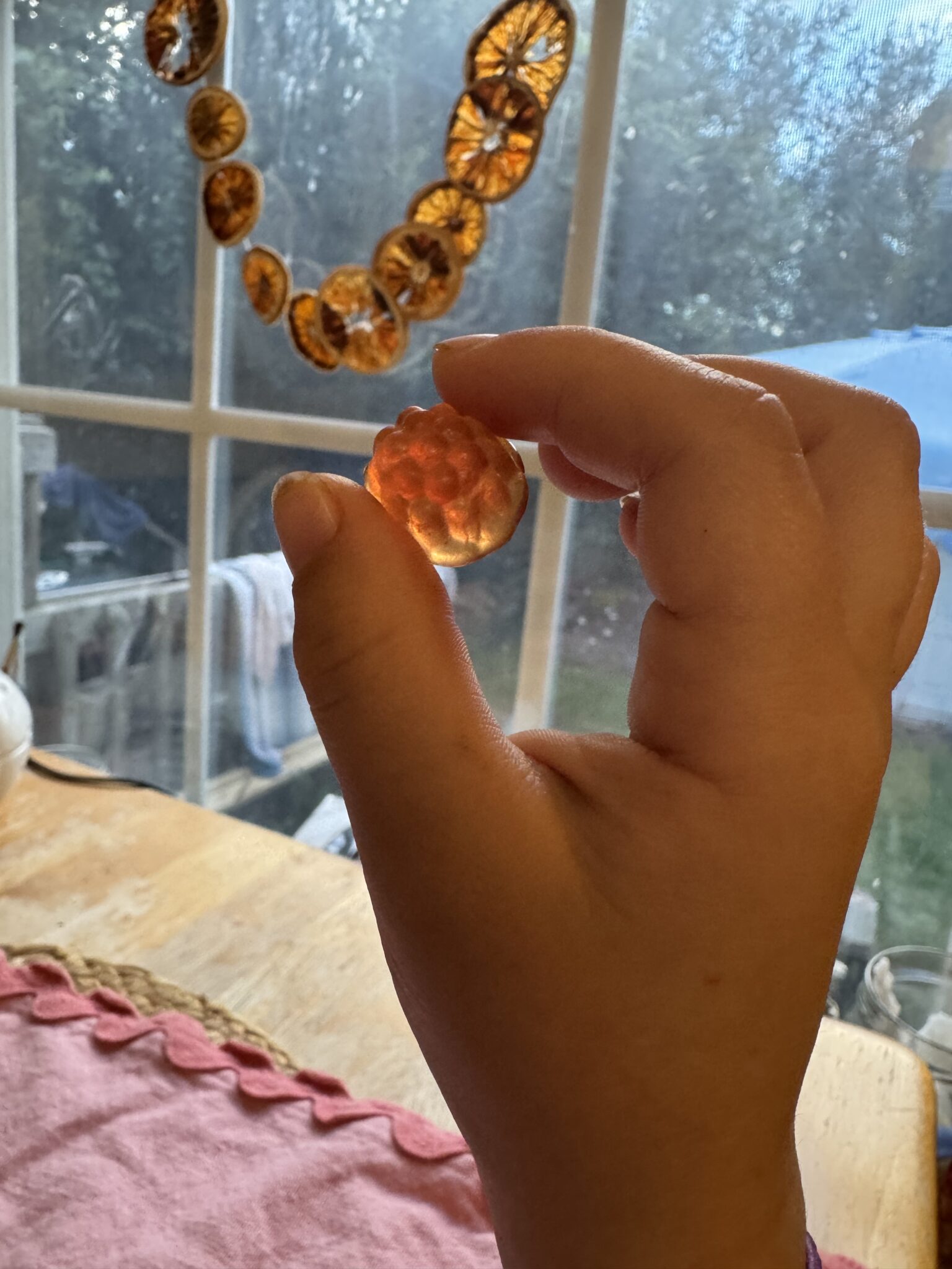 A hand holds up an amber-colored, round object shaped like a berry in front of a window with dried orange slices hanging as decoration.