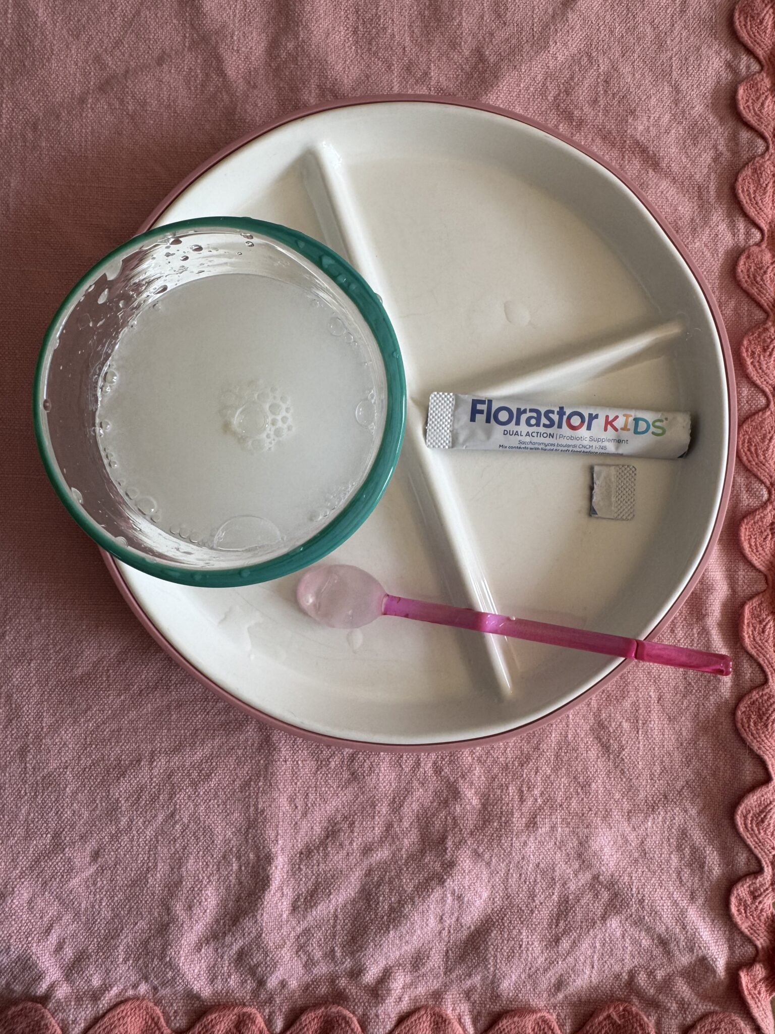 A divided plate with a bowl of cloudy liquid, an empty pink spoon, and a packet labeled "Florastor Kids" on a pink cloth.