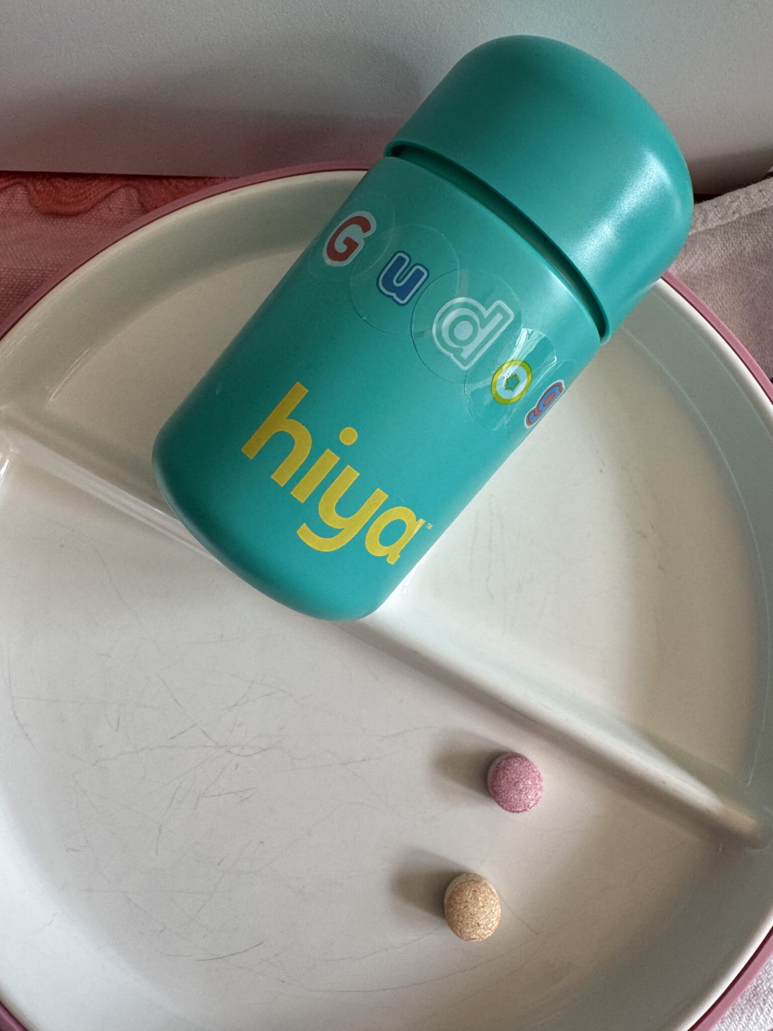 A teal Hiya vitamin container with colorful letters lies on a white divided plate next to one pink and one yellow chewable tablet.
