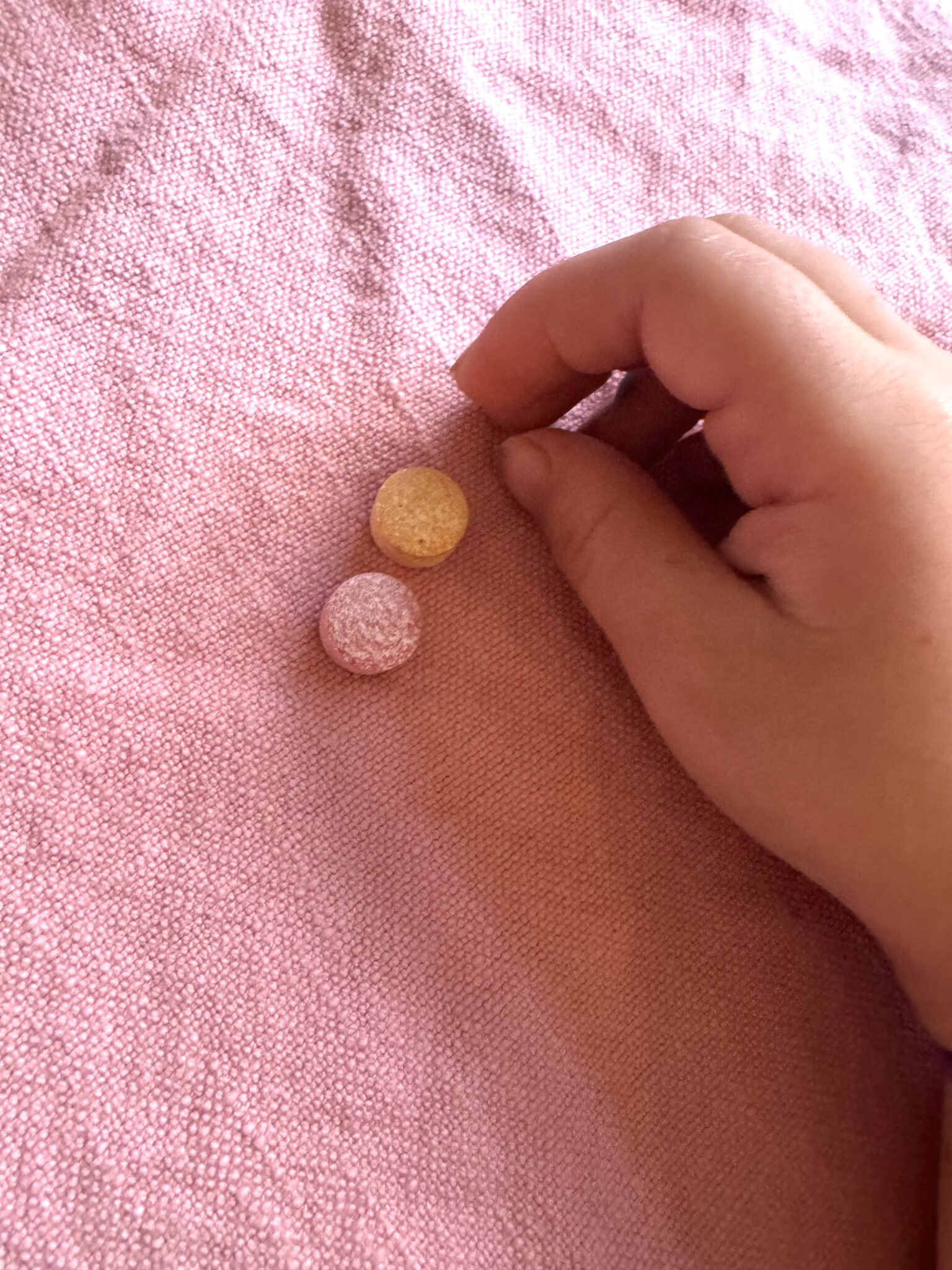A hand reaching toward two round, sugar-coated candies—one yellow and one pink—on a textured pink fabric surface.
