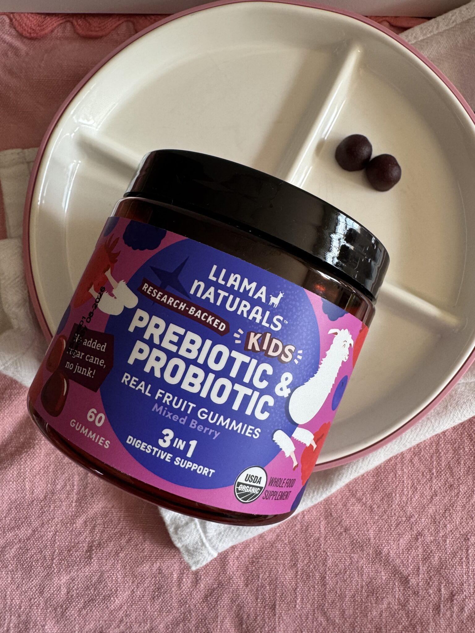 A jar of Llama Naturals Kids Prebiotic & Probiotic Real Fruit Gummies sits on a plate with two gummies beside it, next to a napkin.