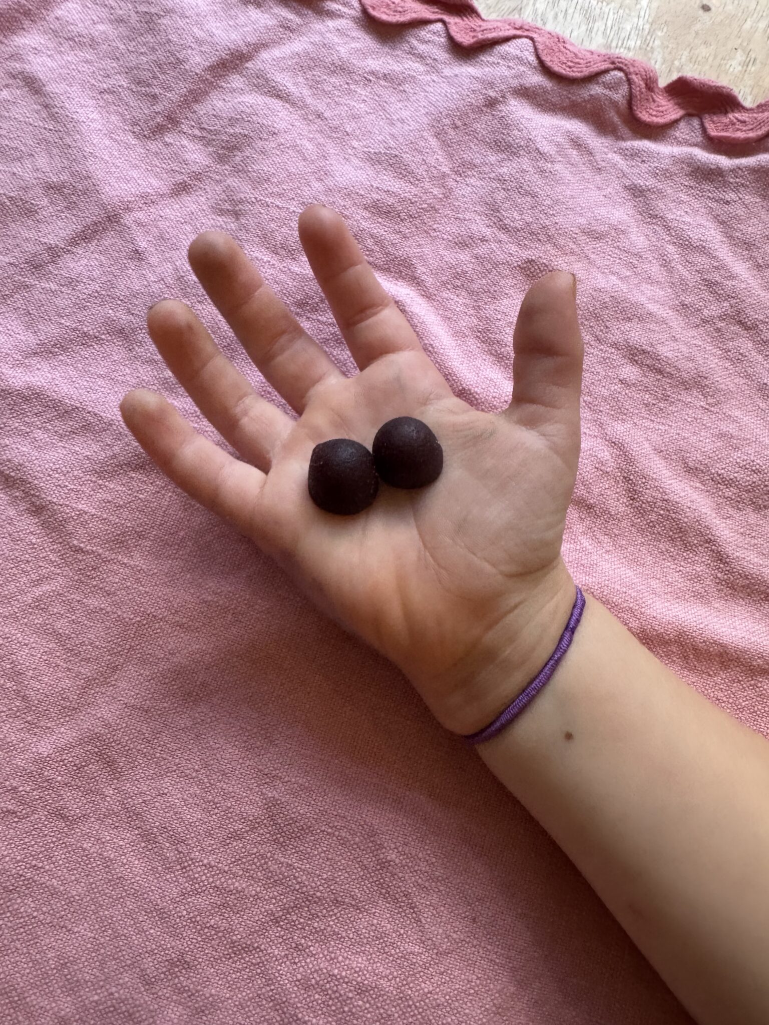 A hand with a purple bracelet holds two small round dark objects, possibly candies or berries, over a pink fabric background.