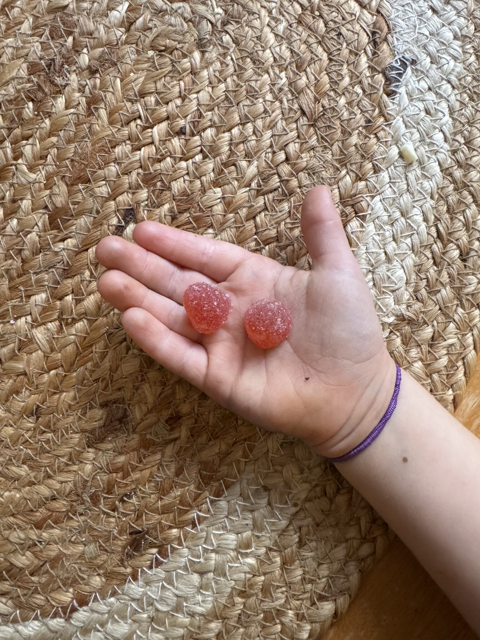 A child's hand holding two round, sugar-coated red gummy candies over a woven mat.