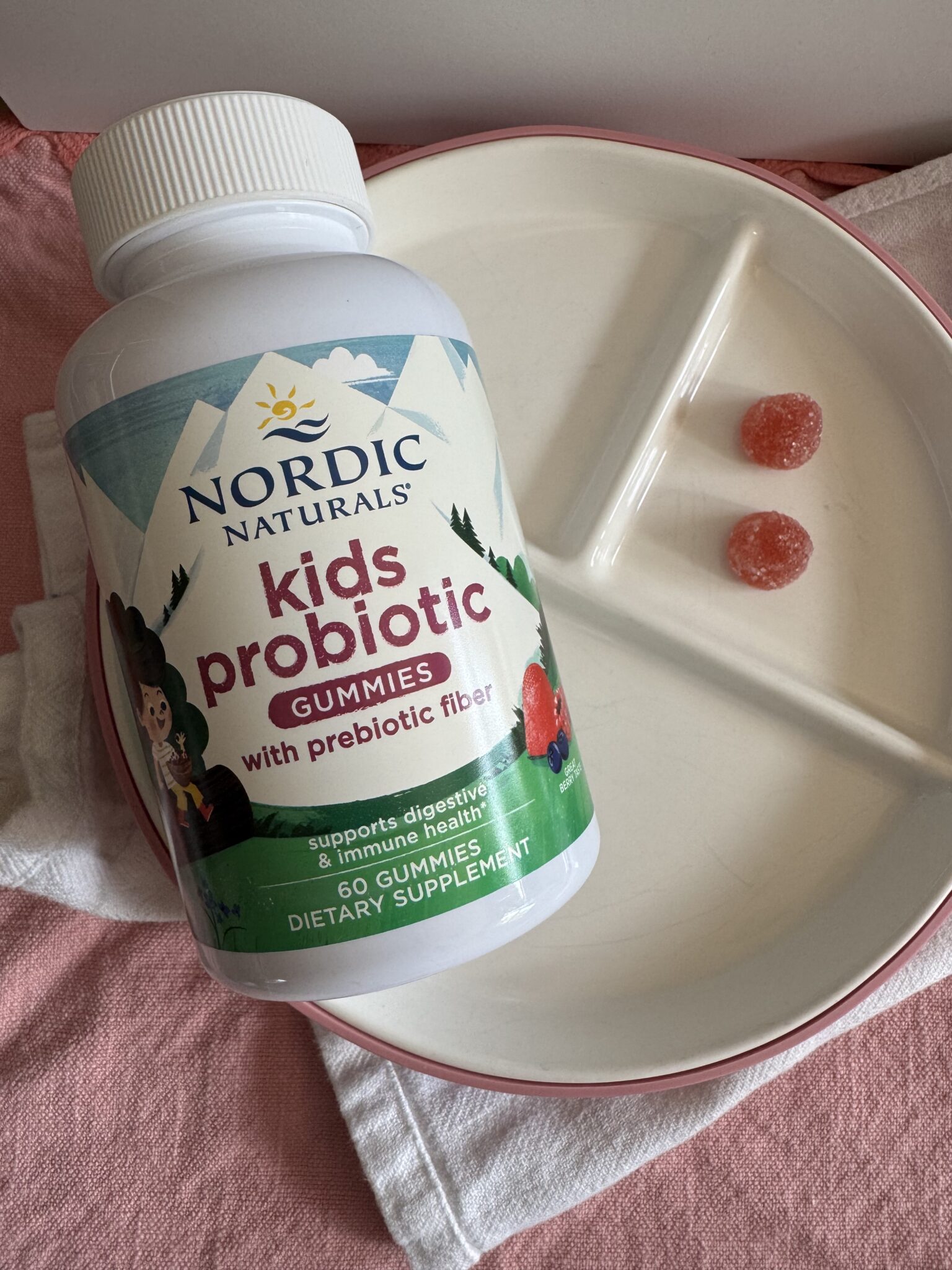 A bottle of Nordic Naturals Kids Probiotic Gummies lies on a plate next to two red gummies.