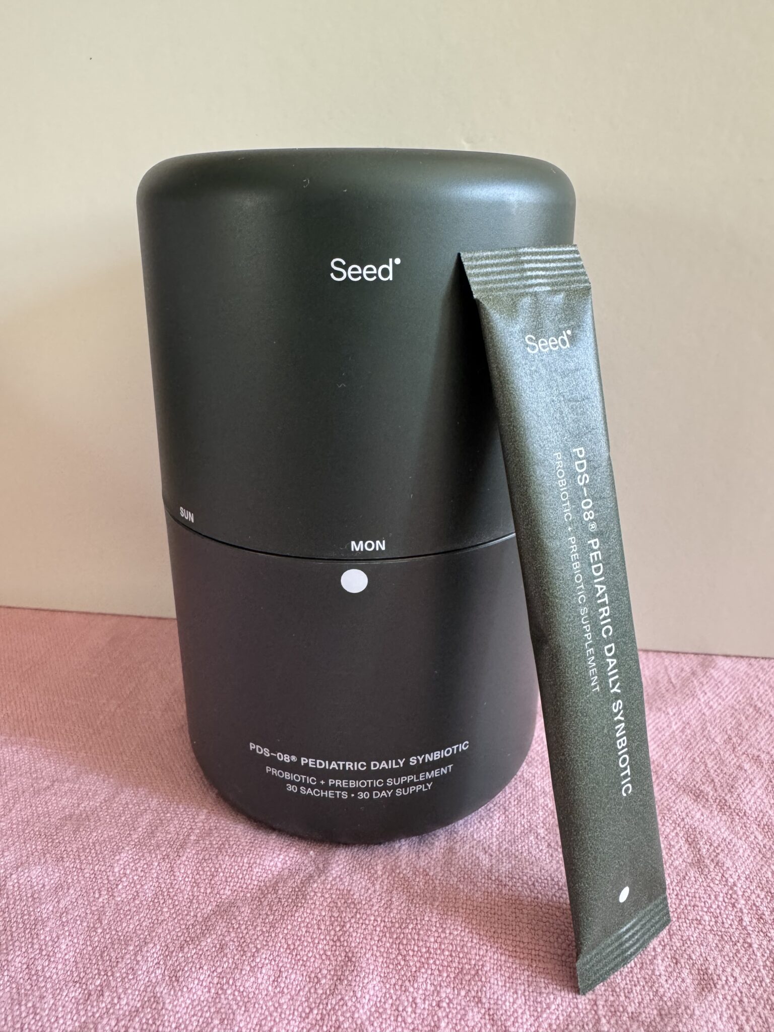 A black Seed container labeled "PDS-08™ Pediatric Daily Synbiotic" stands next to a matching single-use sachet on a pink fabric surface.