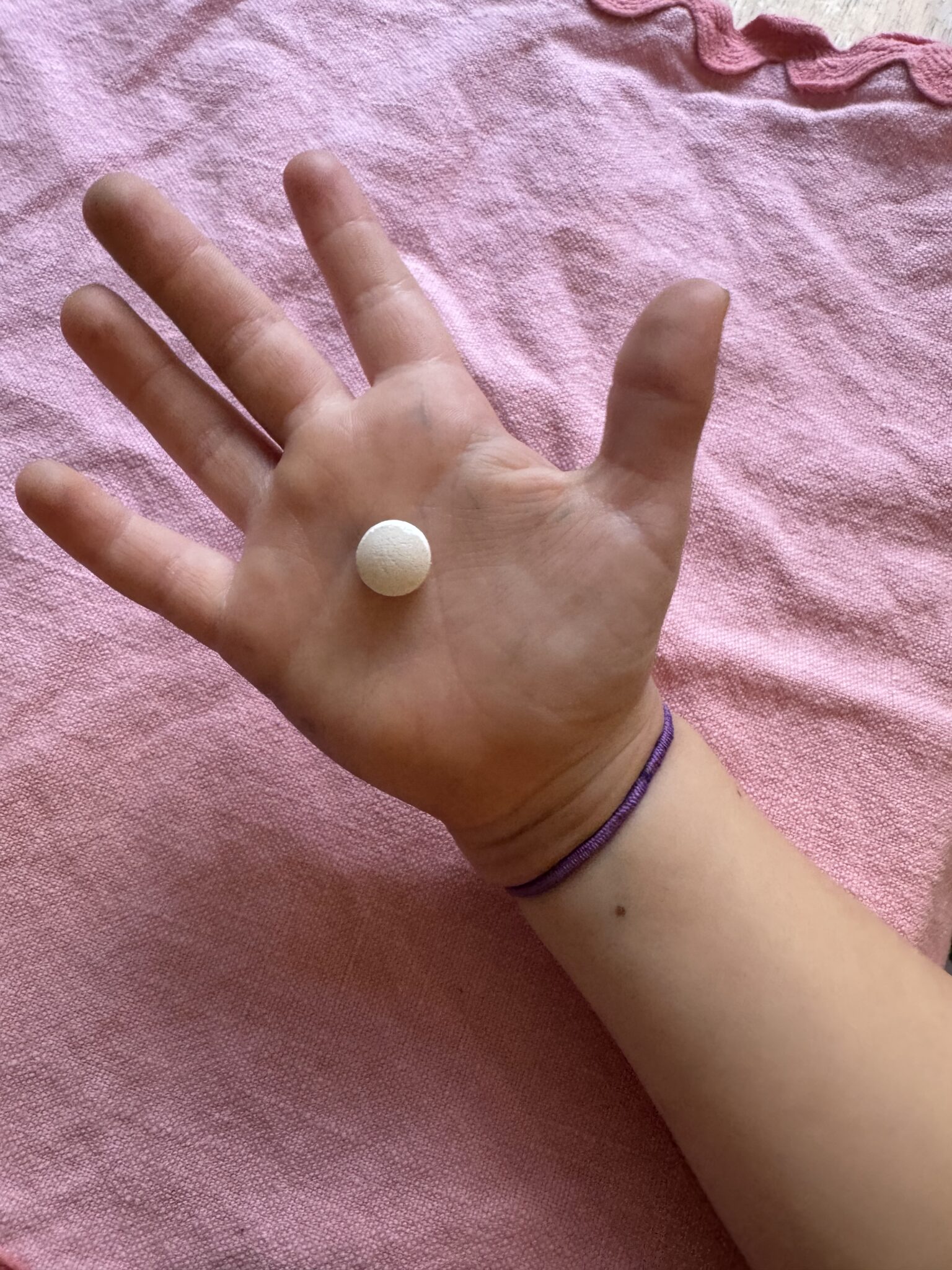 An open hand with a round white pill resting on the palm, over a pink fabric background.