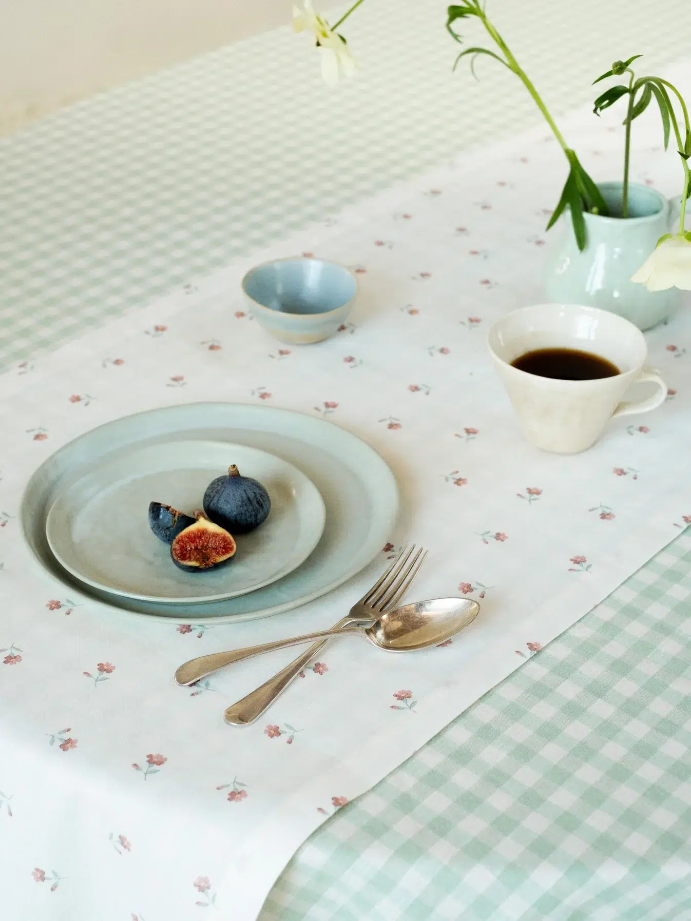 A place setting with a plate holding sliced figs, a cup of coffee, a bowl, cutlery, and vases with flowers on a checkered and floral tablecloth.