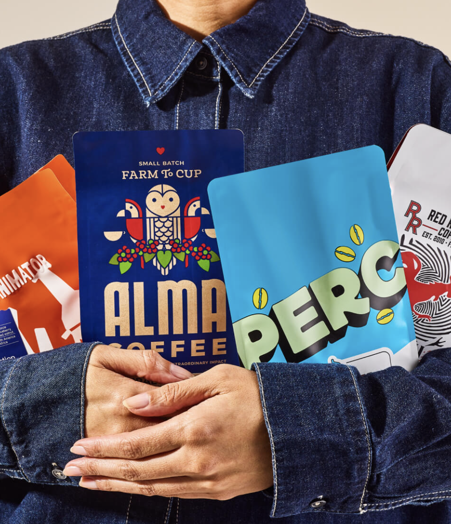 A person in a denim jacket holds several colorful coffee bags with various brand labels and designs visible.