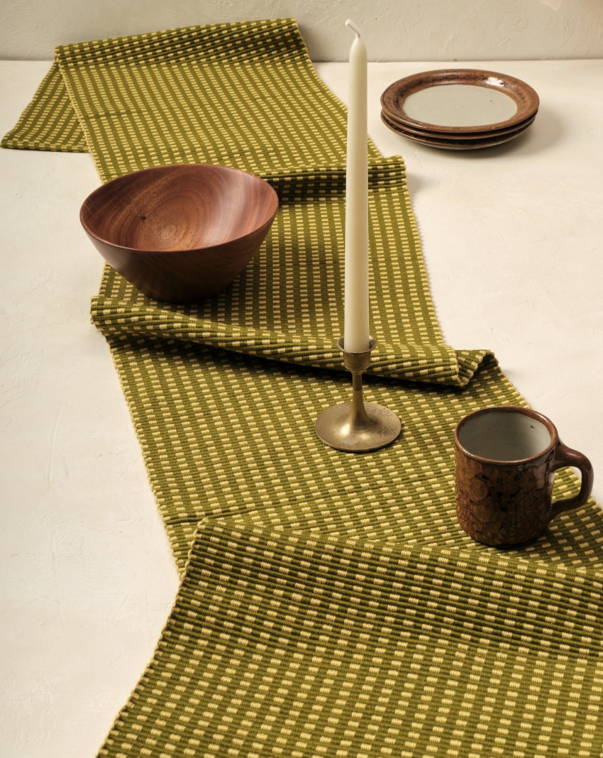 A green patterned table runner with a wooden bowl, brass candlestick with white candle, ceramic mug, and stacked plates on a light surface.