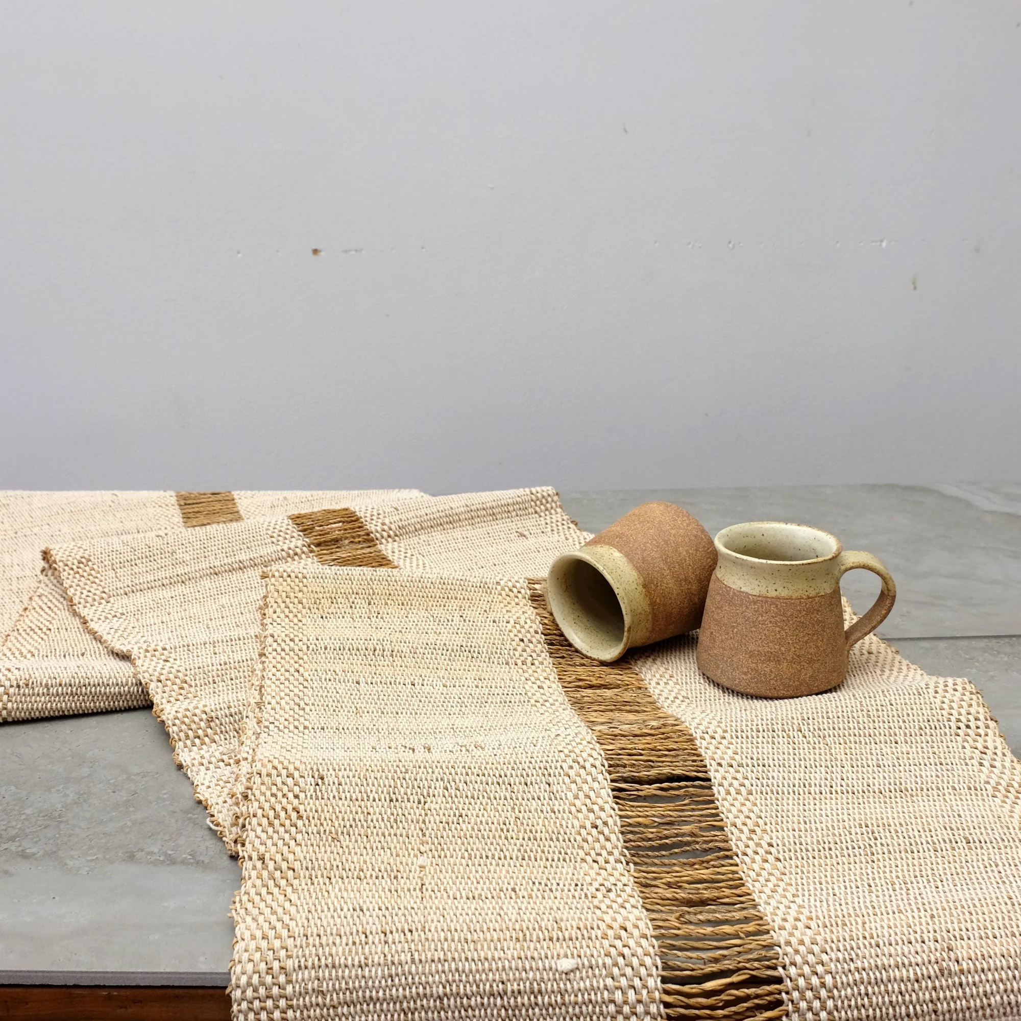 Two ceramic cups sit on a concrete surface beside a woven beige and brown table runner with a textured stripe.