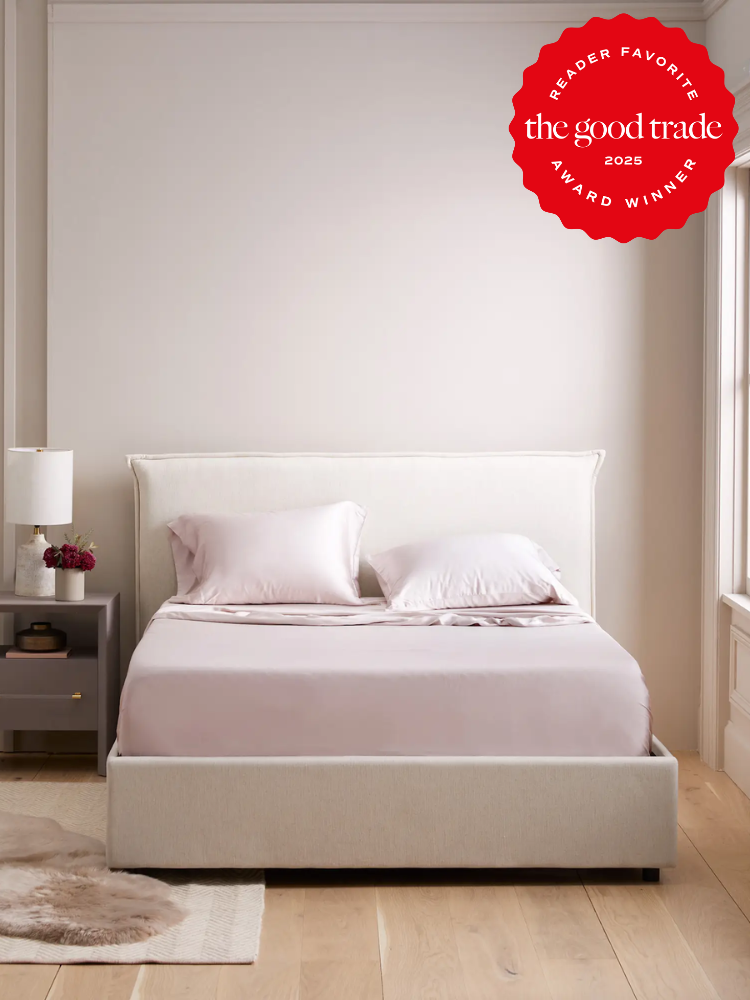 A neatly made bed with light pink sheets and pillows in a minimal bedroom. A nightstand with a lamp is on the left. The top right corner features a "The Good Trade 2025 Award Winner" badge.