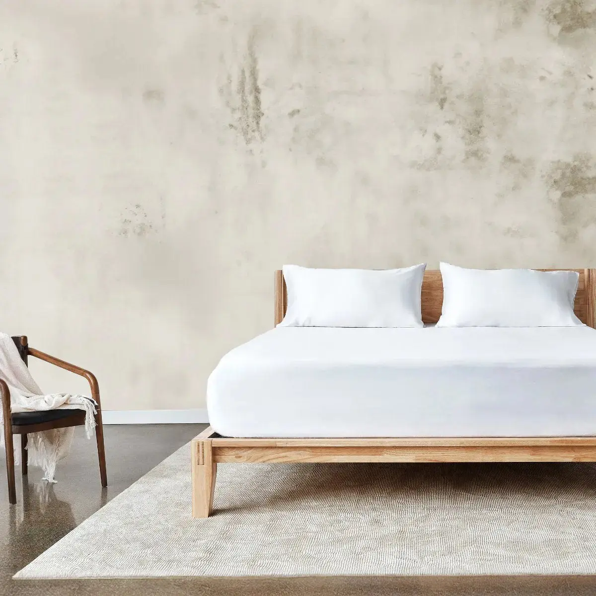 A minimalist bedroom with a wooden bed frame, white bedding, two pillows, a beige rug, a wooden chair with a draped blanket, and a textured off-white wall.