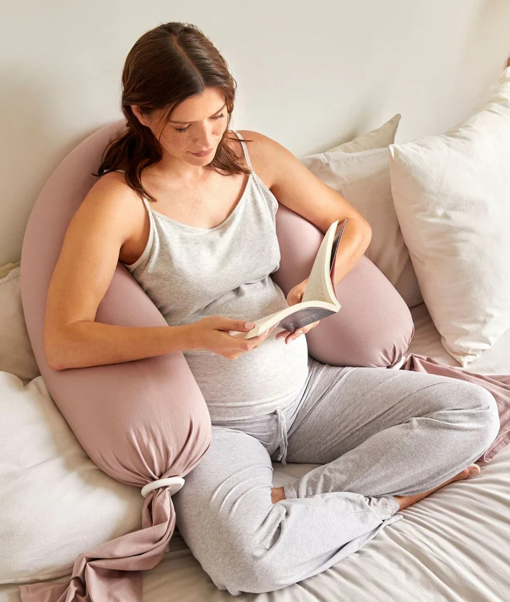 A pregnant woman in loungewear sits on a bed reading a book, supported by a large U-shaped pillow.