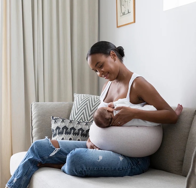 A person sitting on a couch, nursing a baby. They are using a supportive cushion and wearing a white tank top and ripped jeans. The room has a curtain and a framed picture on the wall.