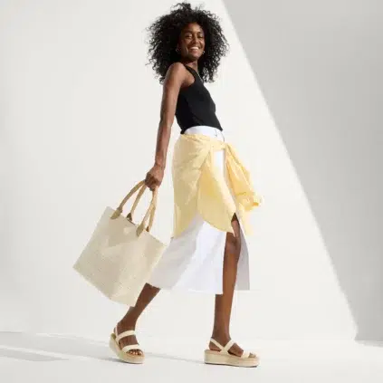 A woman with curly hair wears a black tank top, white skirt, yellow shirt tied at the waist, platform sandals, and carries a large beige tote bag while walking.