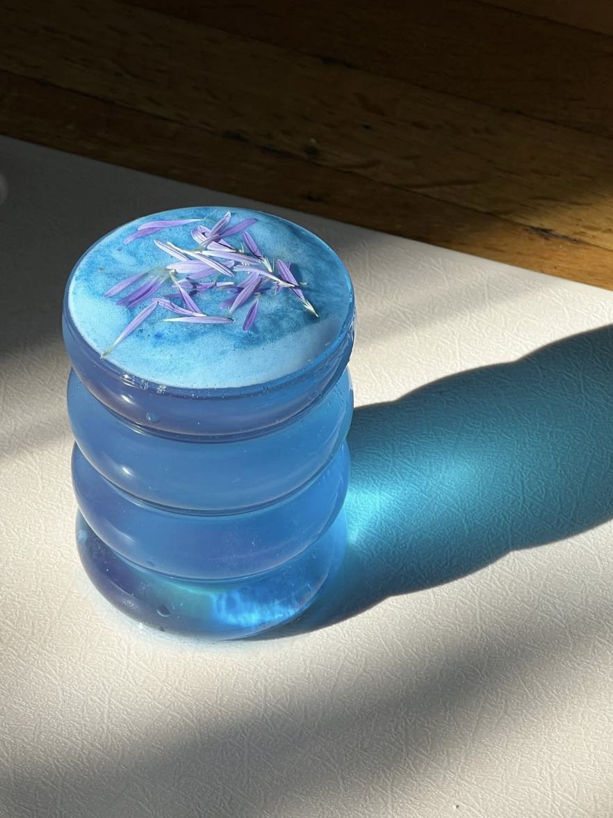 Blue layered candle with purple flower petals on top, casting a shadow on a white textured surface, illuminated by sunlight.