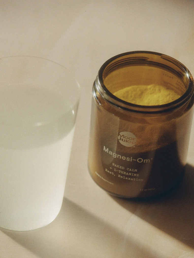 A glass of cloudy liquid beside an open jar labeled "Magnesi-Om" with yellow powder inside, placed on a light surface.