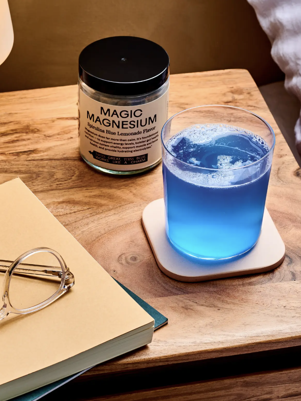 A glass of blue liquid sits on a coaster next to a jar labeled "Magic Magnesium" on a wooden table. Glasses and a closed book are also on the table.