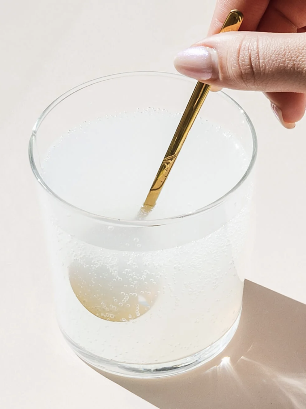 A hand stirs a bubbly white liquid in a clear glass with a golden spoon on a light surface.