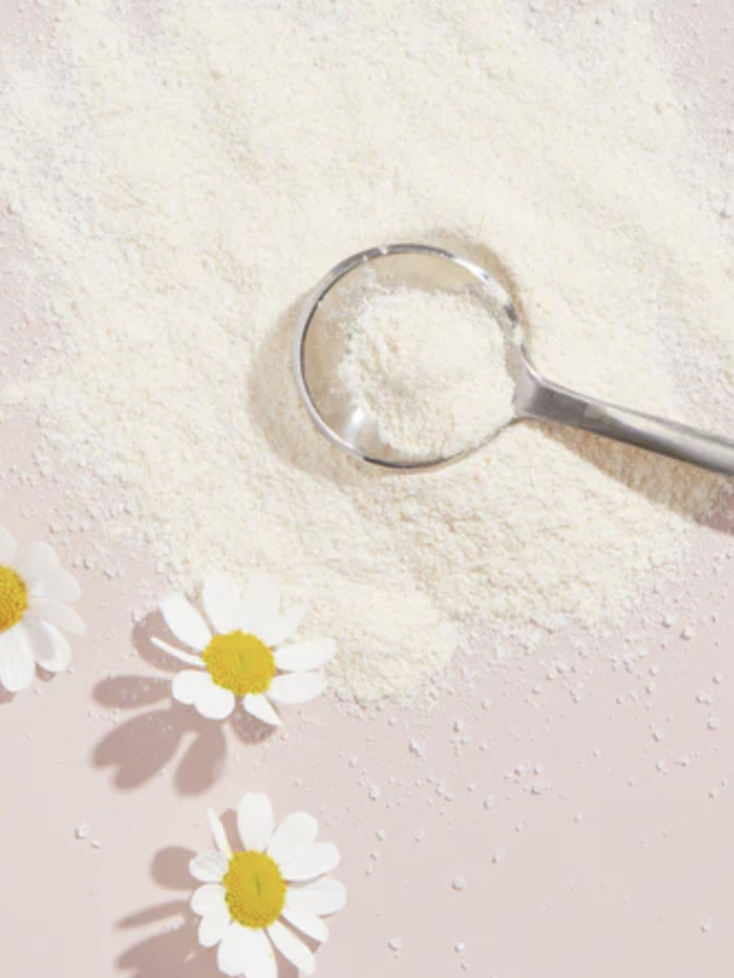 A metal scoop of white powder is on a pink surface with scattered powder and three white daisies.