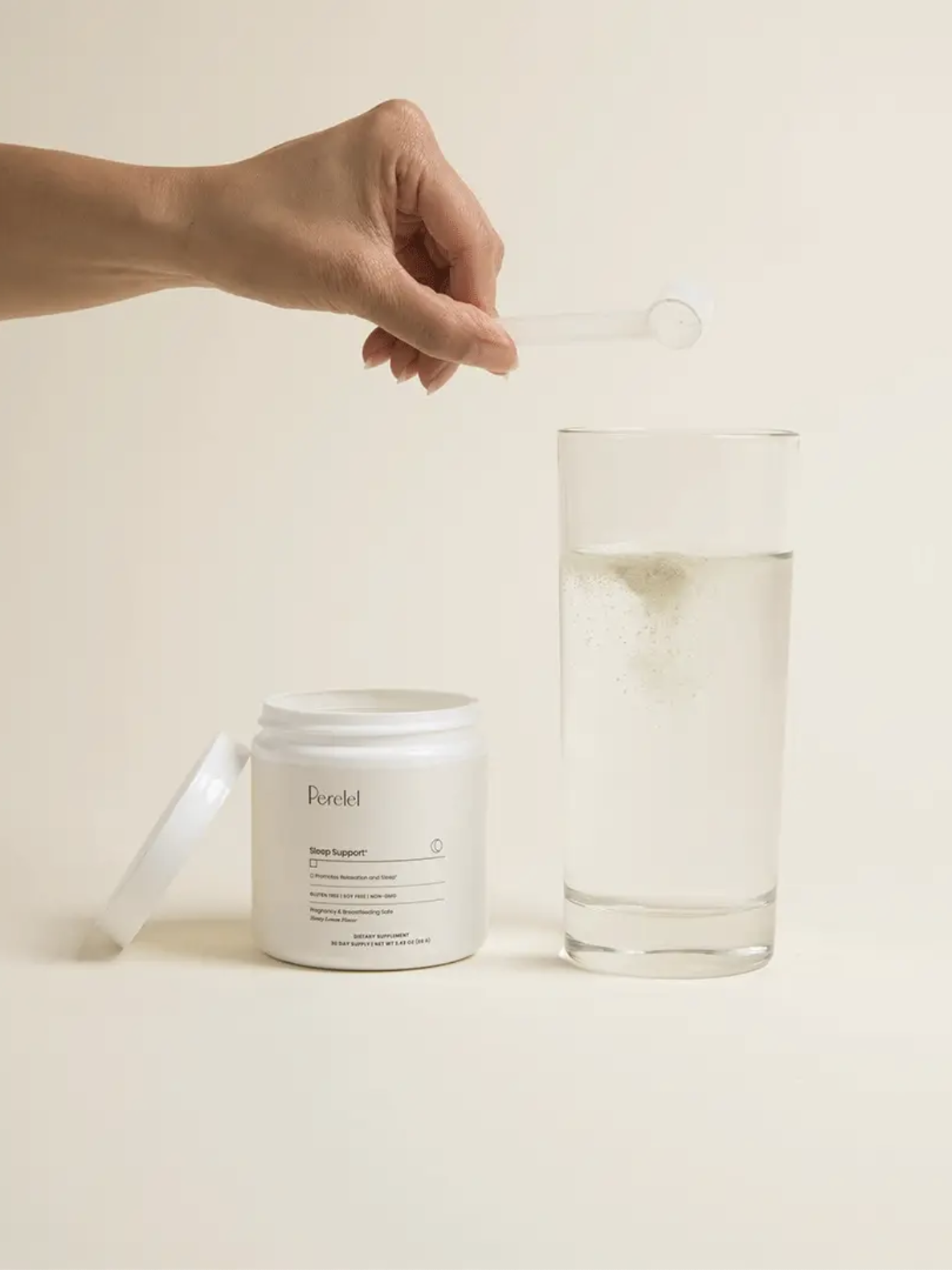 A hand holds a small scoop over an open jar next to a glass of clear liquid on a beige background.